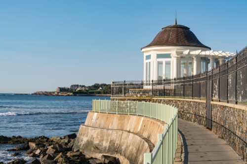 A coastal walkway curves around a stone wall, leading to a white gazebo by the water under a clear blue sky.