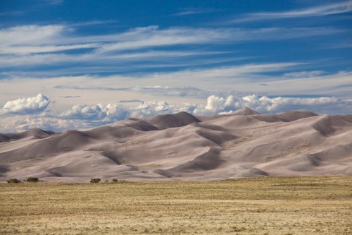 A vast desert landscape with rolling sand dunes under a blue sky adorned with fluffy white clouds.