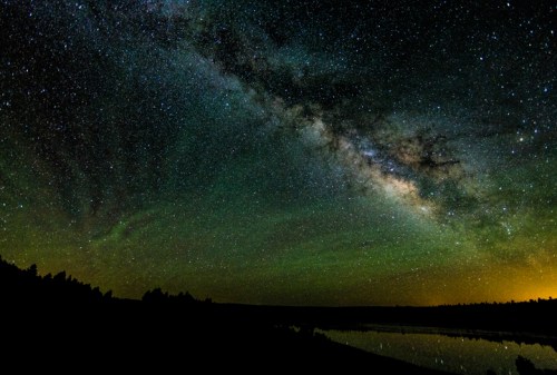 A stunning night sky filled with stars and the Milky Way, reflecting on a calm lake surrounded by dark silhouettes of trees.