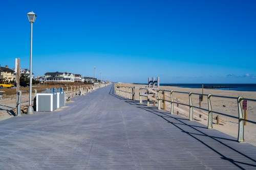 A wide boardwalk stretches along a sandy beach under a clear blue sky, with houses and lampposts in the background.