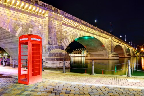 A red telephone booth stands by a river under a lit stone bridge at night, reflecting colorful lights on the water.