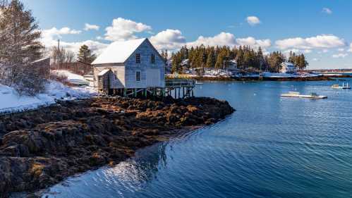 A weathered house on stilts by a calm, snowy shoreline, surrounded by trees and a clear blue sky.