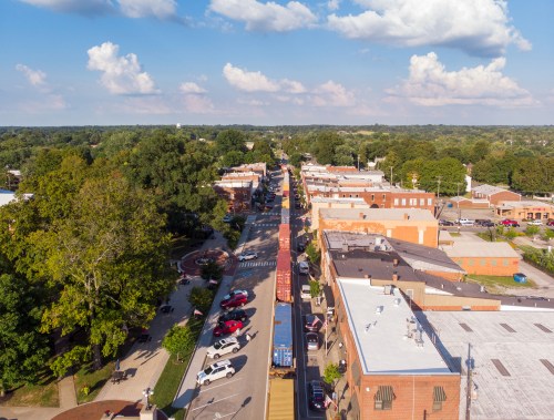 Aerial view of a small town street lined with buildings, trees, and parked cars under a blue sky with clouds.
