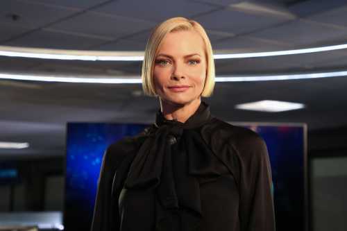 A woman with short blonde hair wearing a black blouse stands confidently in a modern, well-lit studio.