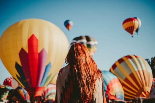 A woman with long red hair stands facing colorful hot air balloons against a clear blue sky.