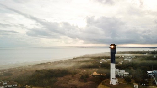 A lighthouse stands tall by the shore, surrounded by mist and a cloudy sky, with the ocean in the background.