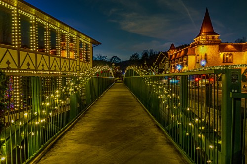 A lit pathway leads through a festive area with buildings adorned in twinkling lights at dusk.