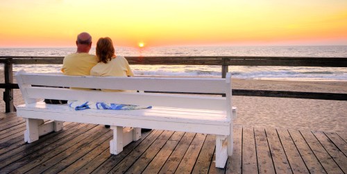 A couple sits on a white bench, watching the sunset over the ocean from a wooden pier.