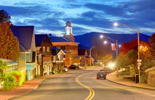 A quiet town street at dusk, lined with shops and trees, with mountains in the background and a clock tower illuminated.