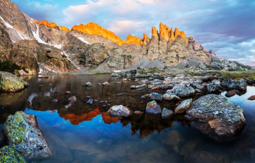Sunlit rocky peaks reflect in a calm lake, surrounded by boulders and lush greenery under a colorful sky.