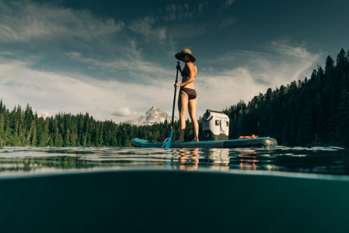 A person stands on a paddleboard in a lake, surrounded by trees and mountains, wearing a hat and swimwear.