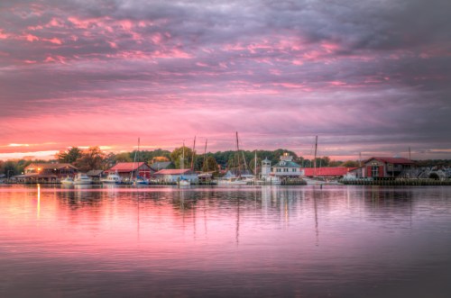 A serene waterfront scene at sunset, with colorful clouds reflecting on calm water and boats docked along the shore.