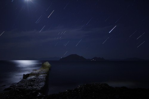 A long exposure shot of a moonlit pier extending into a calm sea, with star trails and distant islands in the background.