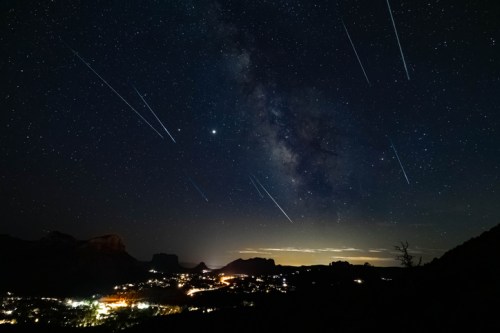 A starry night sky filled with meteors over a lit-up valley and mountains in the distance.