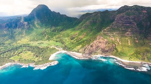 Aerial view of lush green mountains meeting a turquoise coastline with waves crashing on the shore.