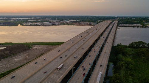 Aerial view of a wide highway over a river, with vehicles traveling and industrial areas visible in the background.