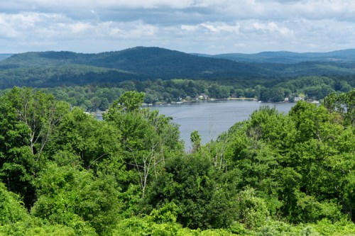 A scenic view of a lake surrounded by lush green trees and rolling hills under a partly cloudy sky.
