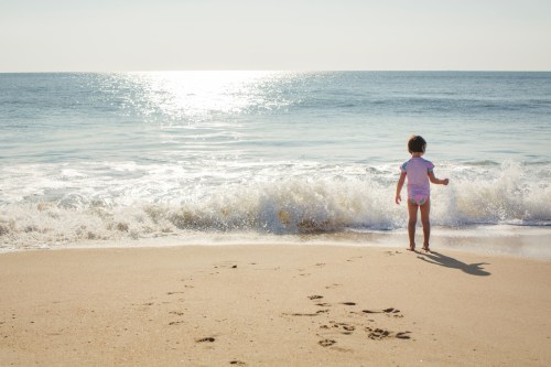 A child stands at the edge of the beach, facing the ocean waves under a bright sky. Footprints lead to the water.