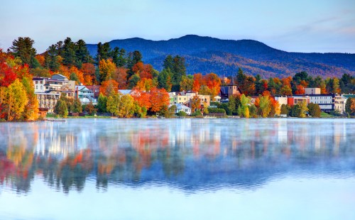 A serene lake reflects colorful autumn trees and a mountain backdrop, with charming buildings along the shore.