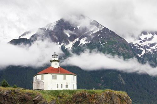 A white lighthouse with a red roof stands against a backdrop of snow-capped mountains and cloudy skies.