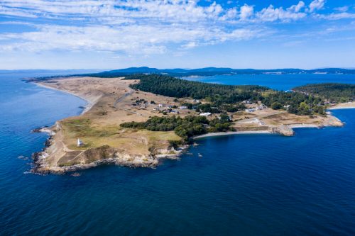 Aerial view of a coastal landscape with a lighthouse, grassy land, and blue waters under a clear sky.