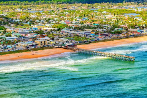 Aerial view of a beach pier with colorful buildings and waves, surrounded by greenery and a coastal town.