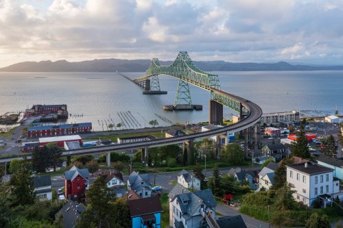 Aerial view of a green bridge spanning water, with a coastal town and mountains in the background under a cloudy sky.