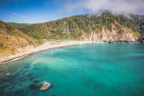 A scenic coastal view featuring turquoise waters, a sandy beach, and lush green hills under a clear blue sky.