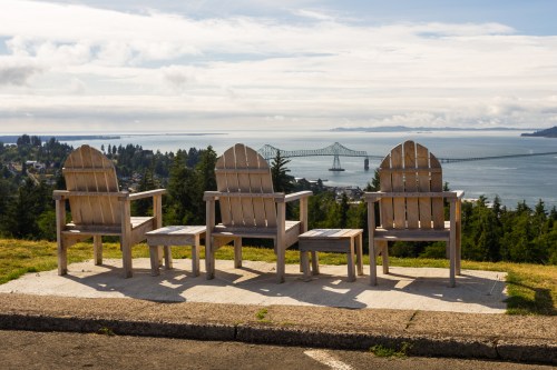 Three wooden chairs overlook a scenic view of a bridge and water, surrounded by trees under a cloudy sky.