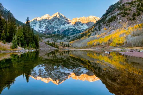 A serene lake reflects majestic mountains and autumn trees under a clear blue sky.