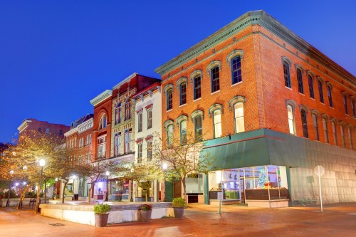 Historic brick buildings line a street, illuminated at dusk with trees and planters in the foreground.