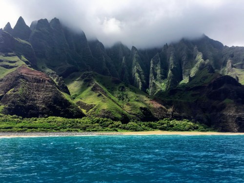 Lush green mountains rise dramatically from the ocean, shrouded in clouds, with a sandy beach in the foreground.