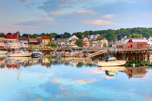 A serene harbor scene with boats and colorful buildings reflecting in calm water under a blue sky.