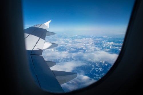 View from an airplane window showing a wing above clouds and snow-capped mountains in the distance.