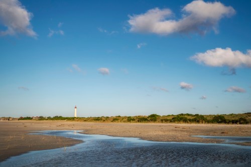 A serene beach scene with a lighthouse in the distance, blue sky, fluffy clouds, and a small tidal pool.