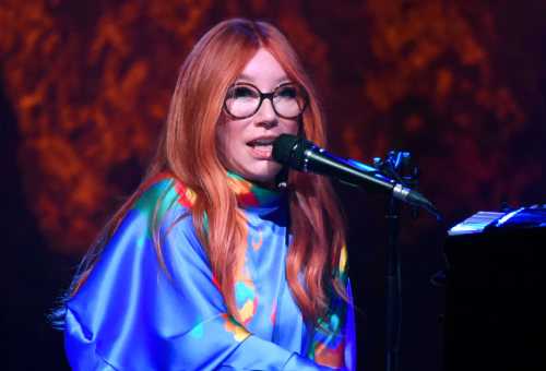 A musician with long red hair and glasses performs at a microphone, wearing a colorful, flowing outfit.