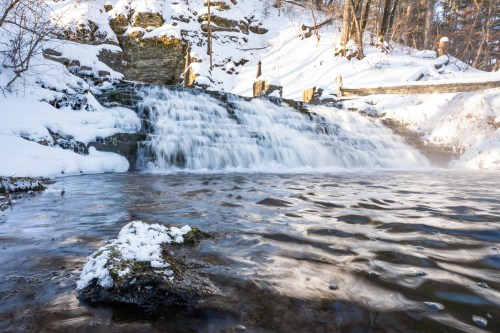 A serene winter scene featuring a waterfall cascading over rocks, surrounded by snow-covered trees and a calm river.