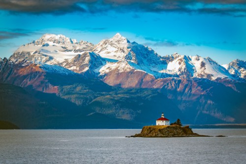 A lighthouse on a small island with snow-capped mountains in the background and calm waters in the foreground.