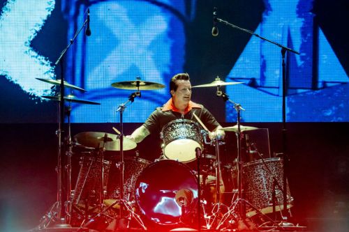 A drummer performs on stage, wearing a colorful shirt, with a leopard-print drum set and vibrant background lights.
