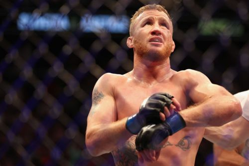 A shirtless MMA fighter with tattoos, wearing gloves, stands in the octagon, looking focused and determined.