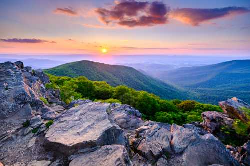 A stunning sunset over rolling green mountains, with rocky foreground and colorful clouds in the sky.