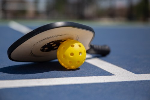 A yellow pickleball resting on a paddle, placed on a blue court with white lines.