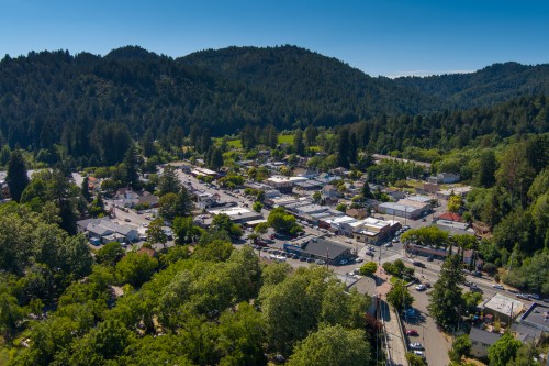 Aerial view of a small town surrounded by lush green hills and trees, with buildings and parked cars visible.