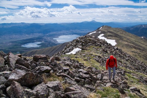 A hiker in a red jacket climbs rocky terrain with a scenic view of mountains and a lake in the background.