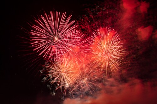 Colorful fireworks burst in the night sky, creating a vibrant display of red and orange against a dark background.