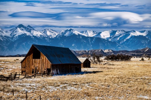 A rustic barn in a snowy field, with majestic mountains and dramatic clouds in the background.