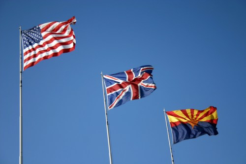 Three flags waving against a clear blue sky: the American flag, the British flag, and the Arizona state flag.