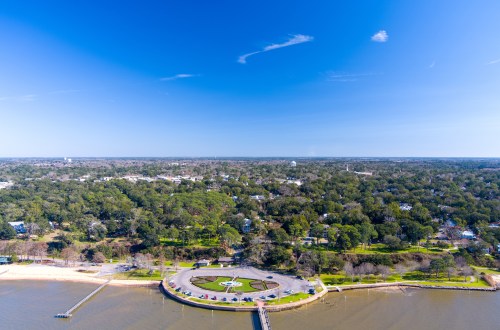 Aerial view of a waterfront park with a circular flower bed, surrounded by trees and a clear blue sky.