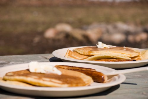 Two plates of pancakes with butter and a sausage link, set on a wooden table outdoors.