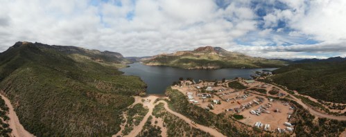 A panoramic view of a lake surrounded by mountains, with a campsite visible along the shore under a cloudy sky.